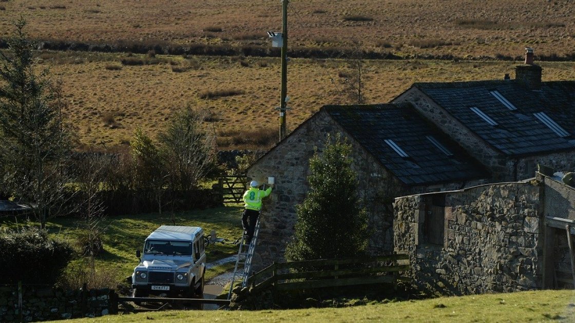 EE engineer on ladder against a farmhouse surrounded by fields in rural environment