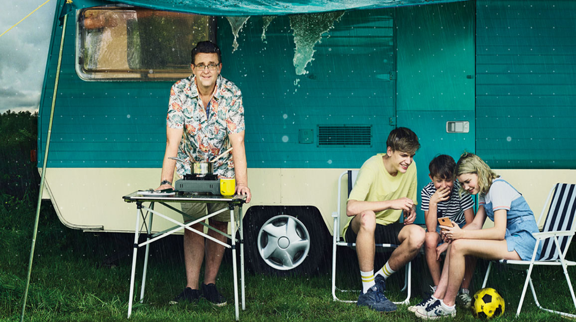 kids huddled round a phone while sitting outside a camper van in the rain, while dad stands by a camp stove